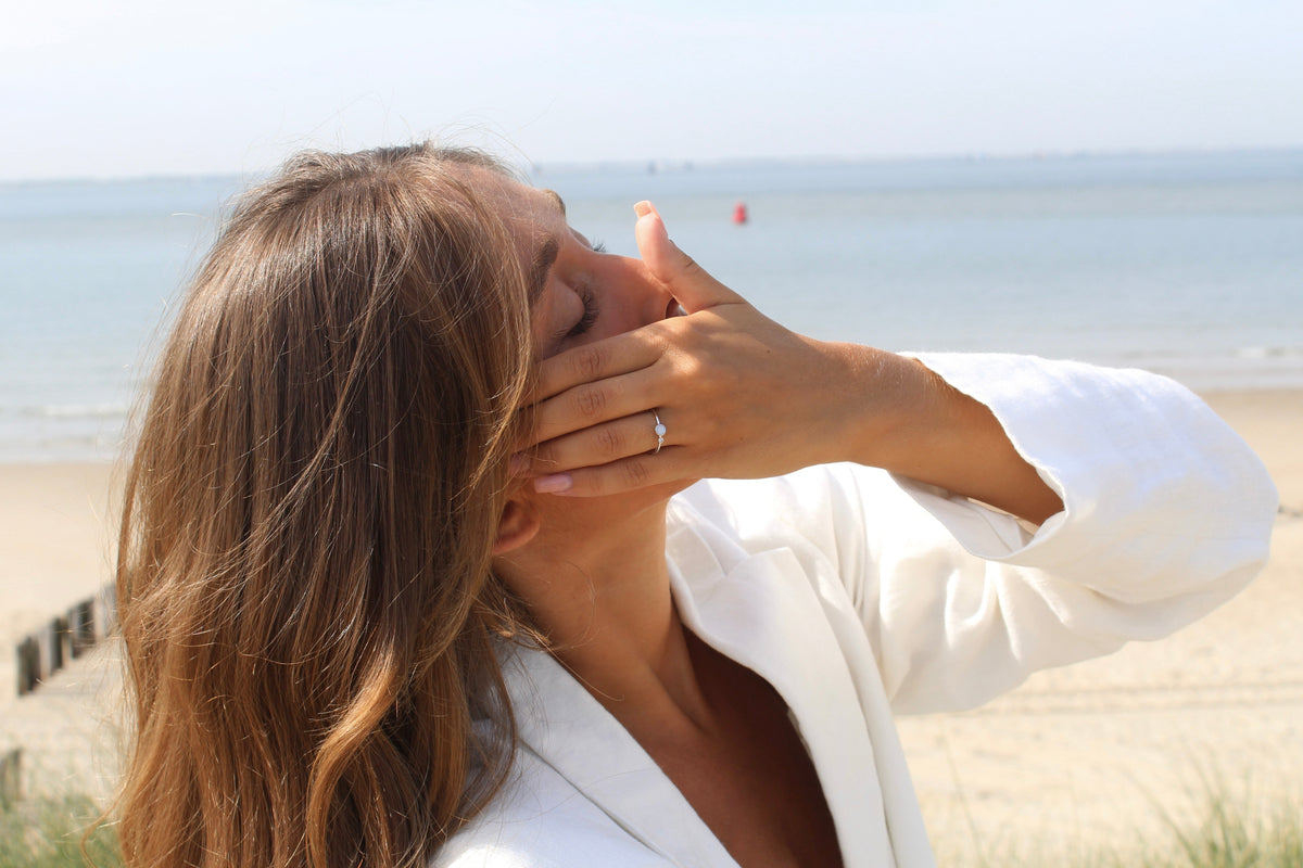 Moedermelk-ring in massief witgoud met een ronde zirkonia, gedragen door een vrouw met een lichte huid en golvend haar, op een strandachtergrond.