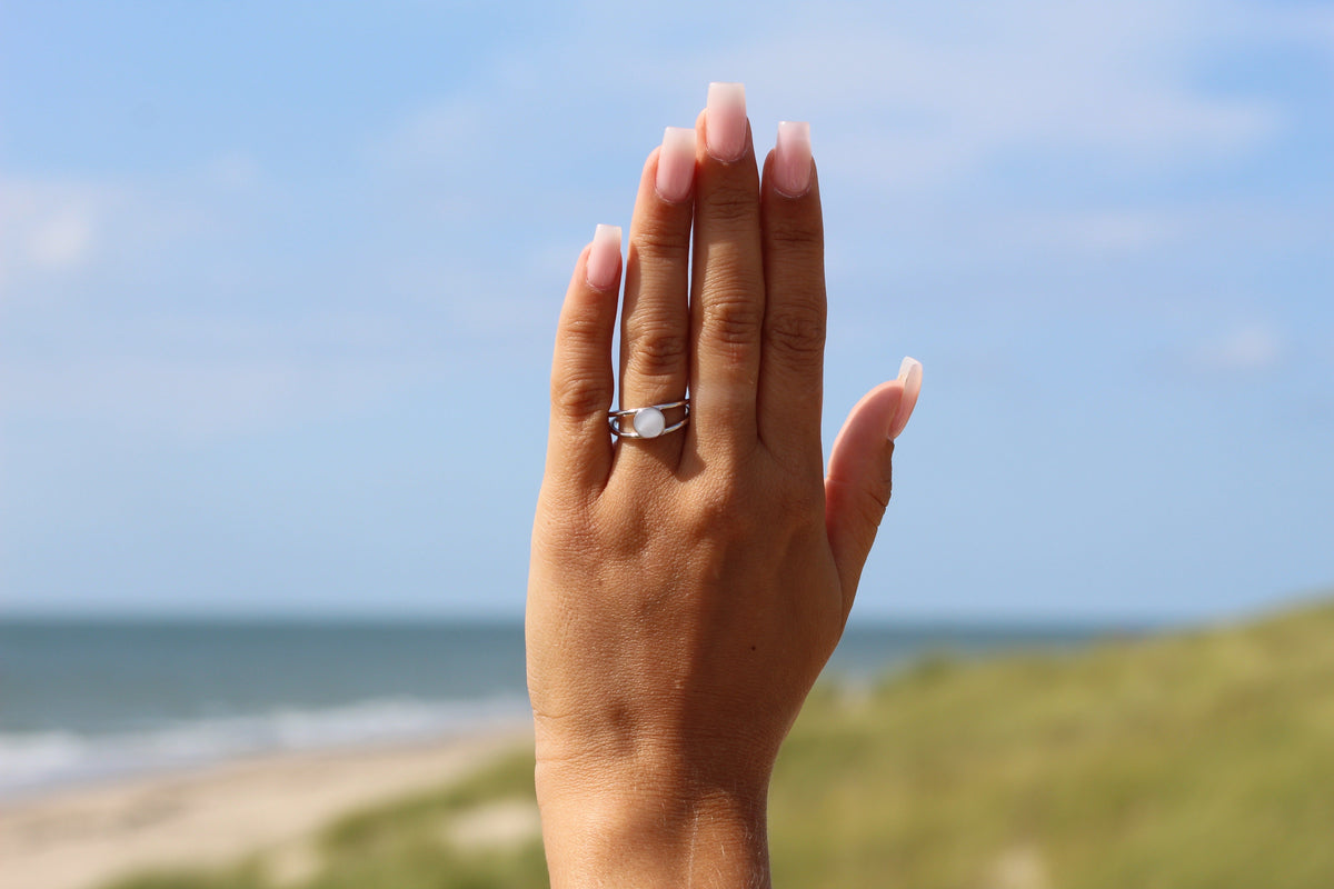 Moedermelkring van 925 zilver met een heldere harssteen, gedragen aan een hand met een natuurlijke manicure, tegen een strandachtergrond.