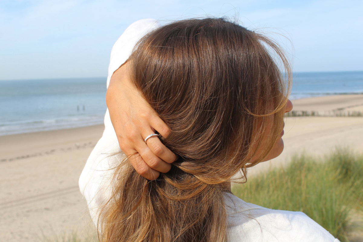 Eenvoudige ring van 925 zilver, gedragen door een vrouw met lang bruin haar, met een natuurlijke achtergrond van zee en strand.