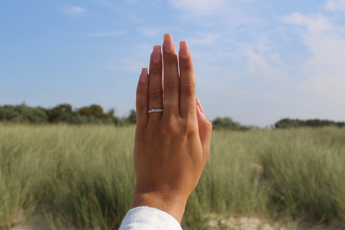 Elegante ring van 925 zilver met een subtiele moedermelkhars, gedragen op een hand met lange, verzorgde nagels, tegen een achtergrond van gras en blauwe lucht.