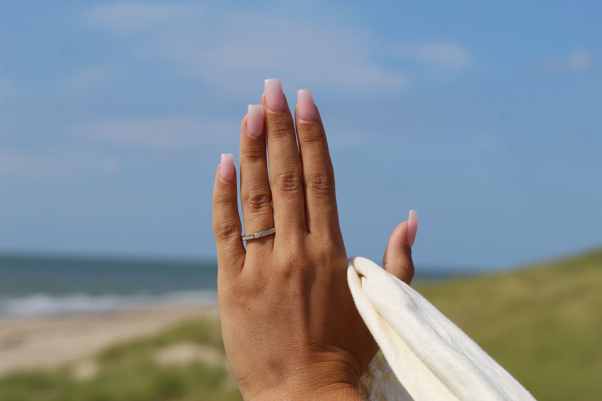 Massief gouden ring met een heldere steen, gedragen op een hand met lange, lichtroze nagels, tegen een strandachtergrond.