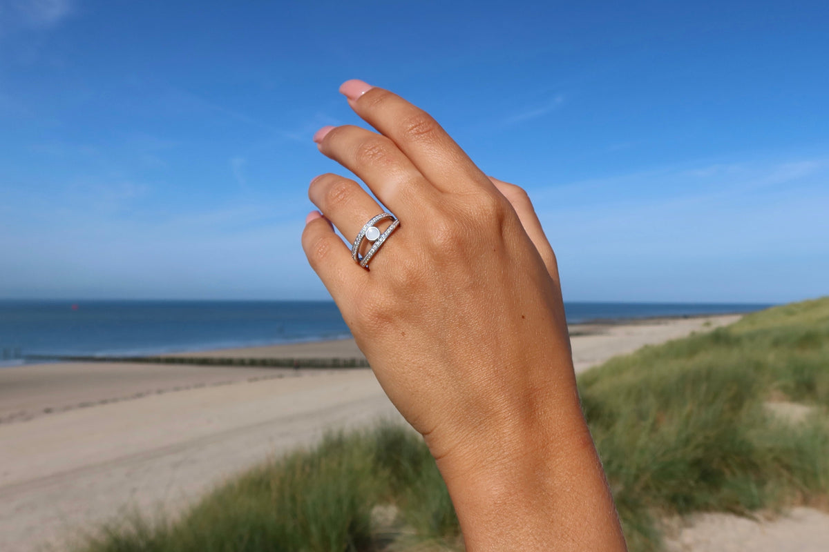 Roségouden moedermelk-ring met een heldere harssteen, gedragen op een hand met een zee- en strandachtergrond.