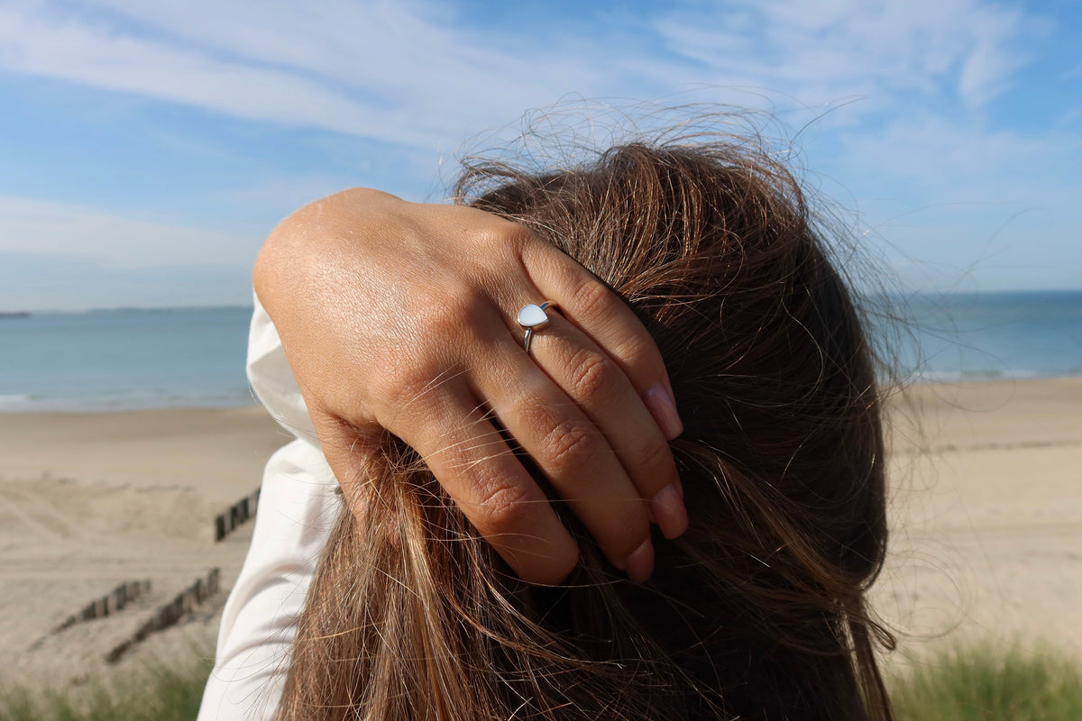 Ring van massief roségoud met een druppelvormige moedermelkhars, gedragen door een persoon met los haar, op een strandachtergrond.