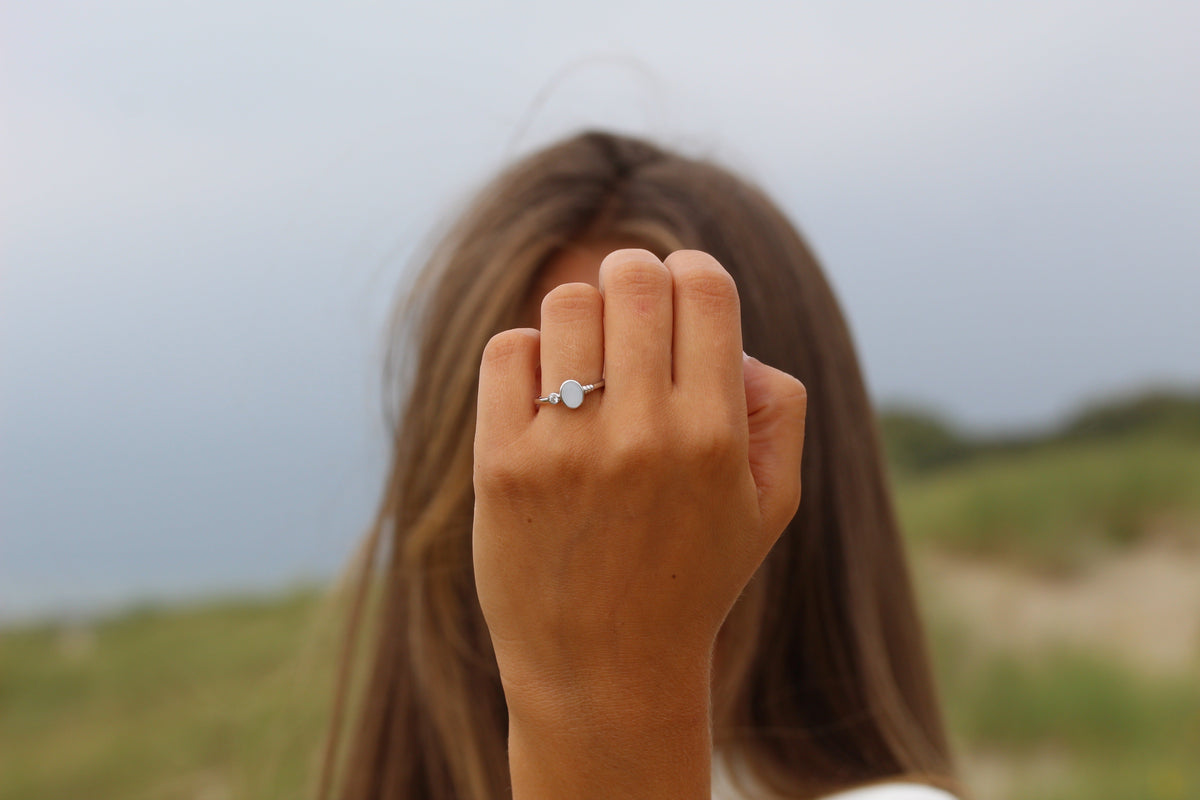 Massief witgouden ring met een ronde moedermelkhars en een kleine zirkonia, gedragen door een persoon met een hand omhoog.