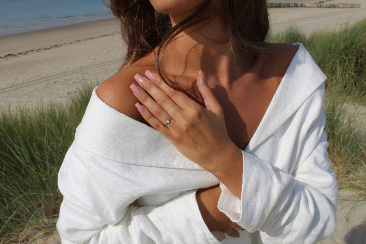 Moedermelk-ring van 925 zilver met een heldere zirkonia, gedragen door een persoon met een blootgestelde schouder, op een strandlocatie.