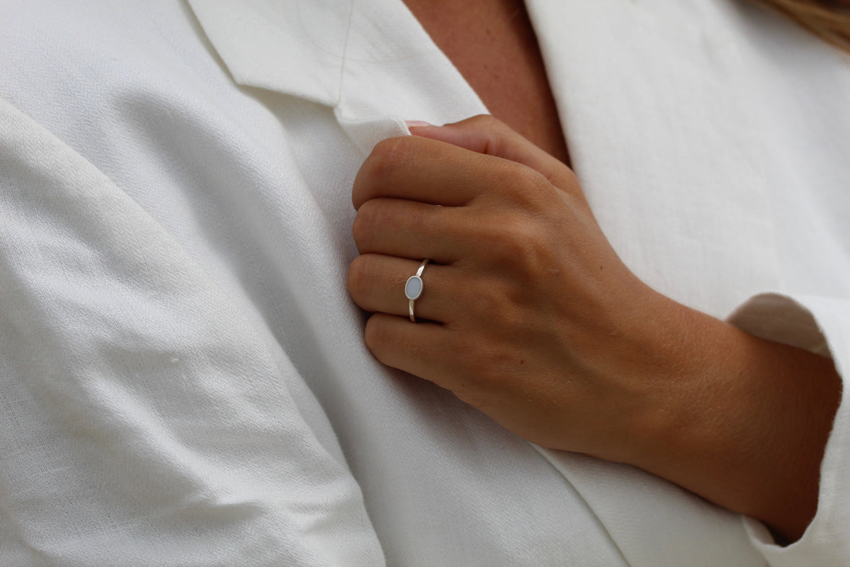 Zilveren ring met een ovale moedermelkharssteen, gedragen op een hand die een witte blazer vasthoudt.