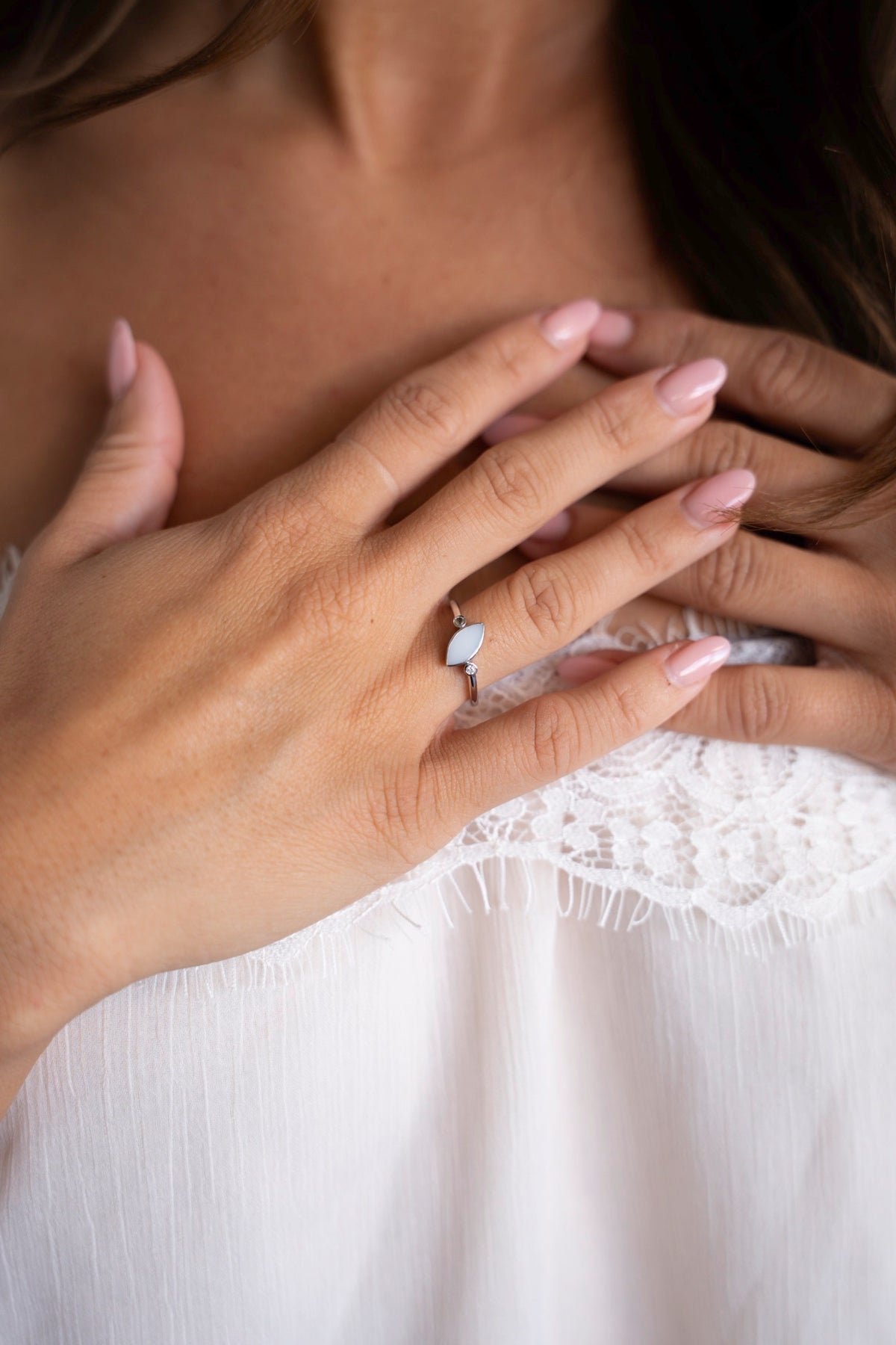 Handen op de borst met de zilveren Lina Duo-ring, een moedermelksteen met twee kleine steentjes, tegen een witte kanten top.