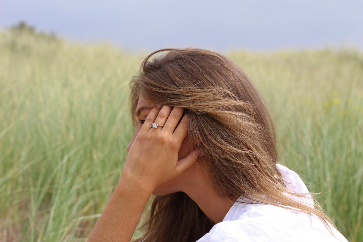 Zilveren moedermelk-ring met een heldere steen, gedragen door een vrouw met los haar, op een achtergrond van gras.