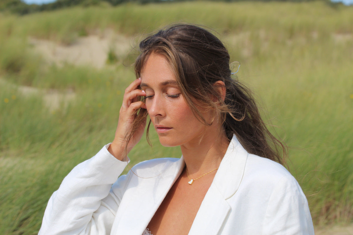 Zilveren moedermelk-hanger in de vorm van een druppel, gedragen door een vrouw in een witte blazer, met een natuurlijke achtergrond van gras.