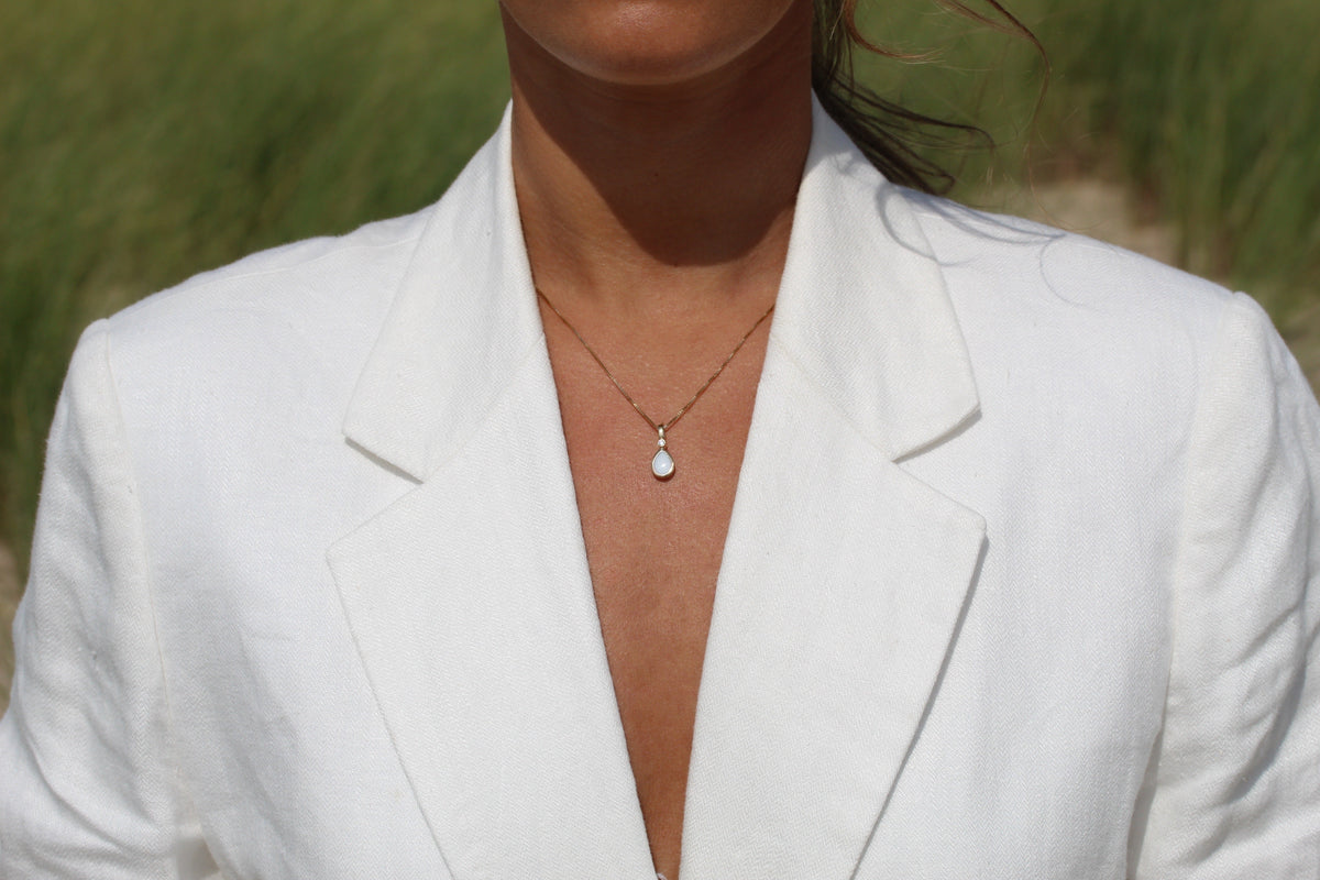 Hanger van massief geelgoud met een druppelvormige moedermelkhars en een subtiele gouden sluiting, gedragen door een persoon in een witte blazer.