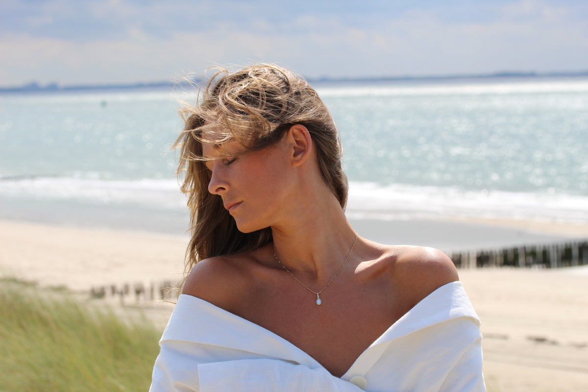 Hanger van massief witgoud met een druppelvormige moedermelkhars, gedragen door een vrouw met een open schoudertop, tegen een strandachtergrond.