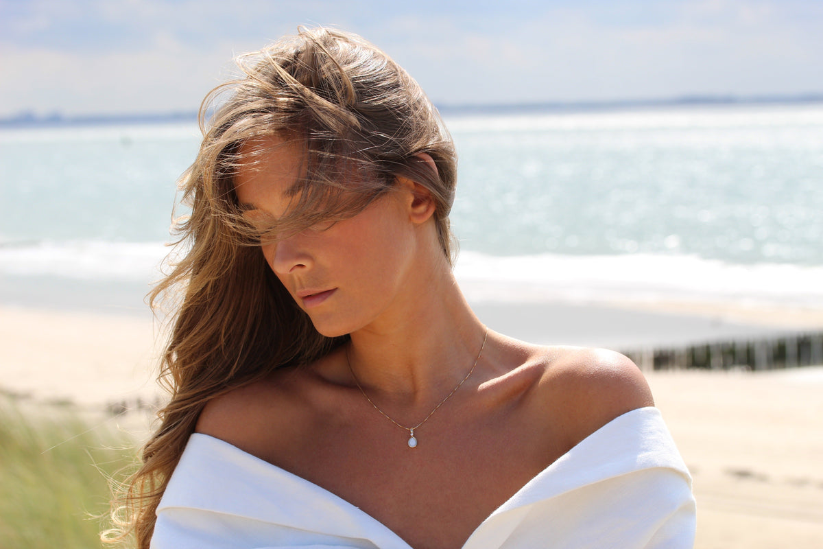Hanger in massief roségoud met een druppelvormige moedermelkhars, gedragen door een vrouw met los haar, tegen een strandachtergrond.