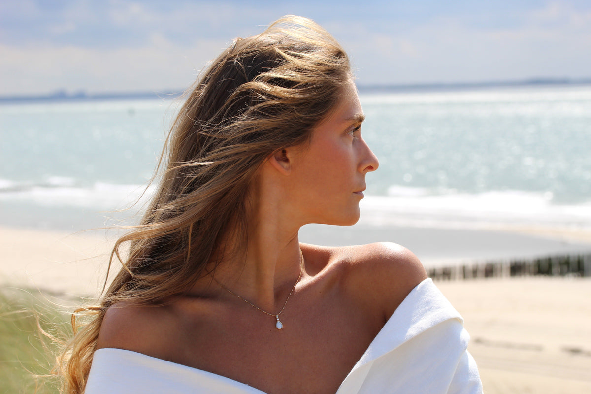 Hanger van massief goud met een druppelvormige moedermelkhars en een subtiele glans, gedragen door een vrouw met los haar aan het strand.