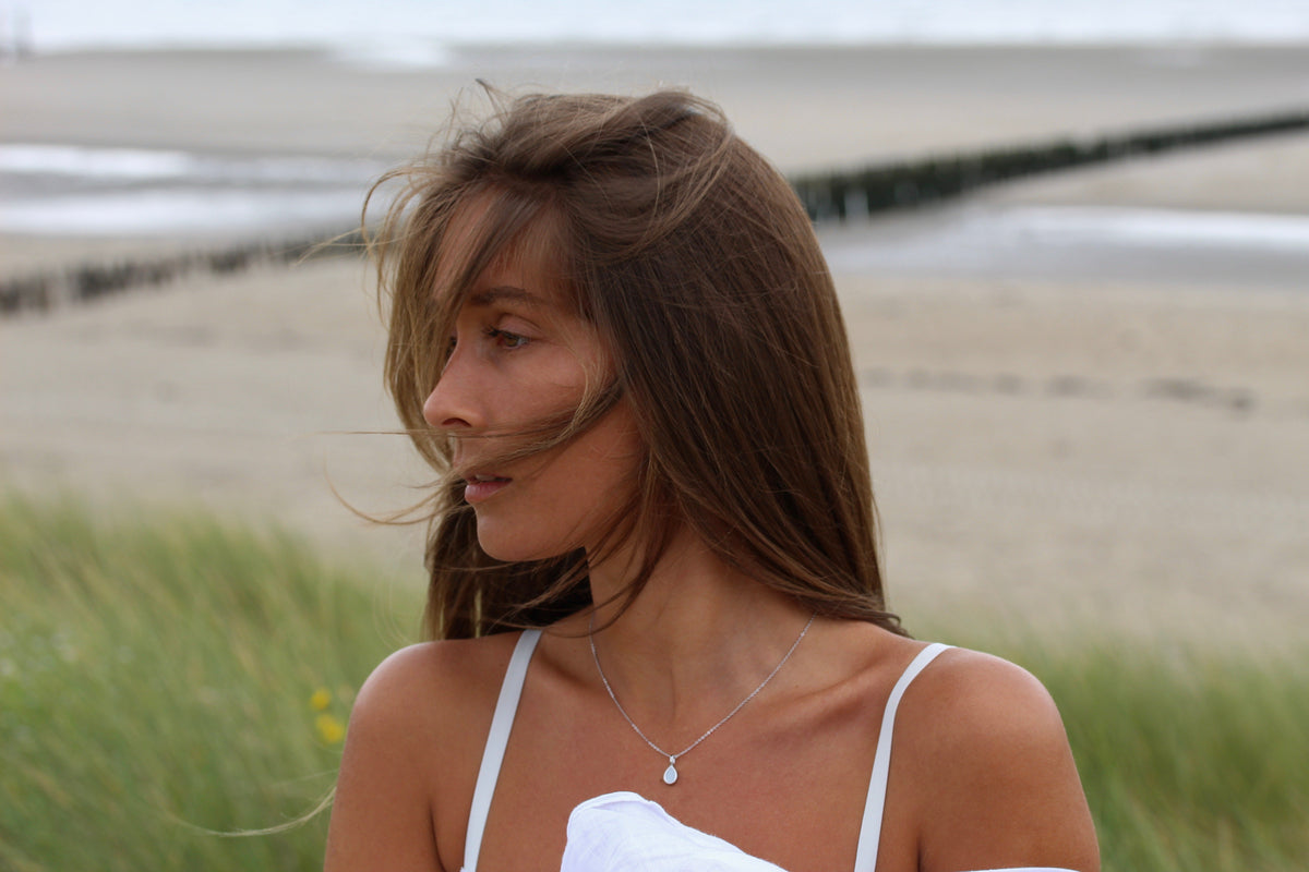 Moedermelk-hanger van 925 zilver in druppelvorm, gedragen door een vrouw met lang bruin haar, op een strandachtergrond.