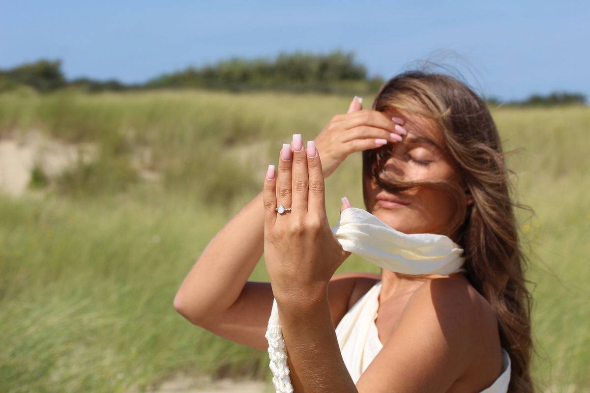 Massief geelgouden moedermelk-ring met een ronde, glanzende harssteen, gedragen door een vrouw met een lichte huid en golvend haar, in een natuurlijke omgeving.