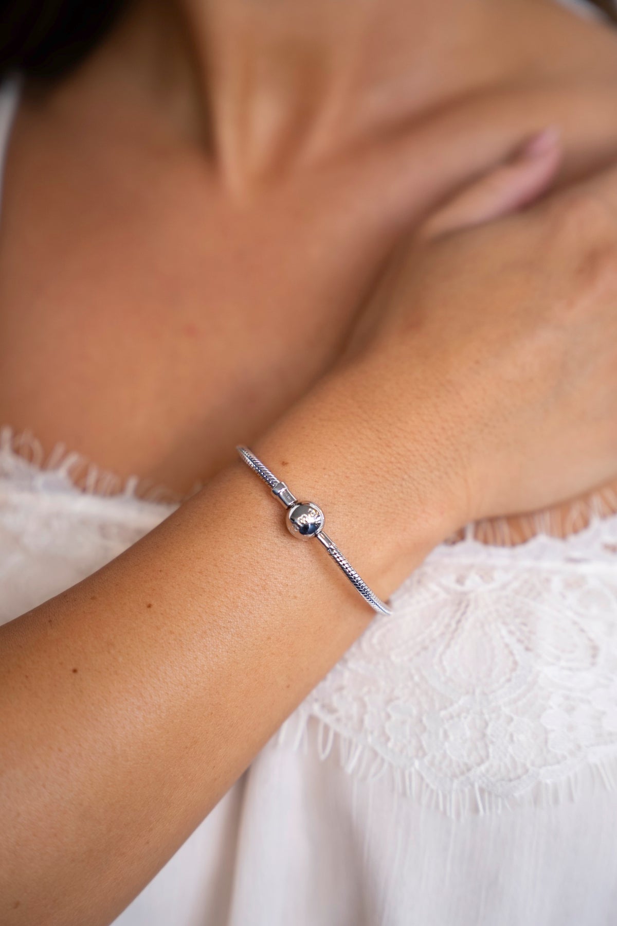 Zilveren bedelarmband om de pols, de hand rust op de borst boven een witte kanten top.